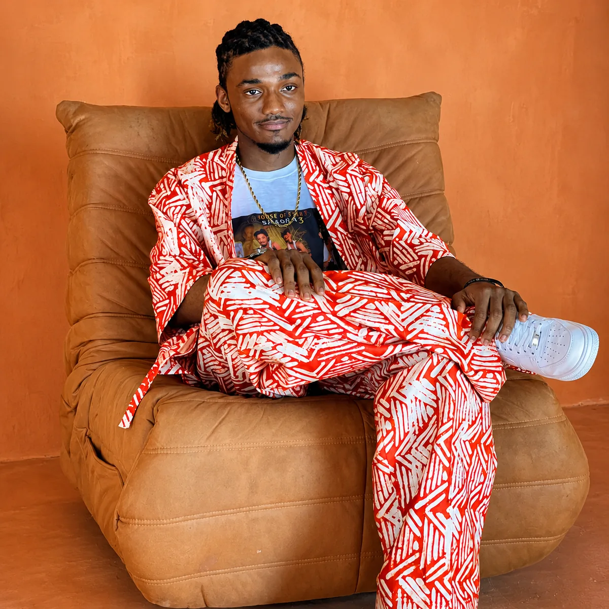 Joseph Sesay seated on a tan couch in a red-and-white patterned traditional outfit with House of Stars Season 3 tee